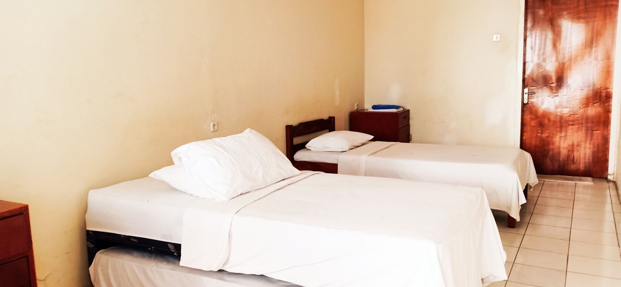 A room with two single beds, each with white bedding and a wooden headboard. There is a small bedside table with a lamp on one side and a wall socket above the bed. The room has a tiled floor and a wooden door on the right.