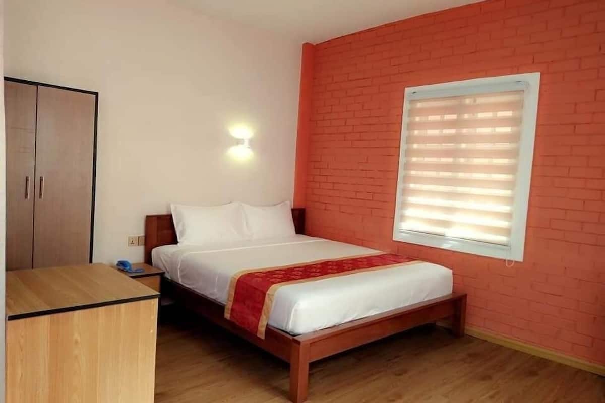 A single-bed room with a wooden bed, a wardrobe, a wall-mounted light, and a window with blinds.