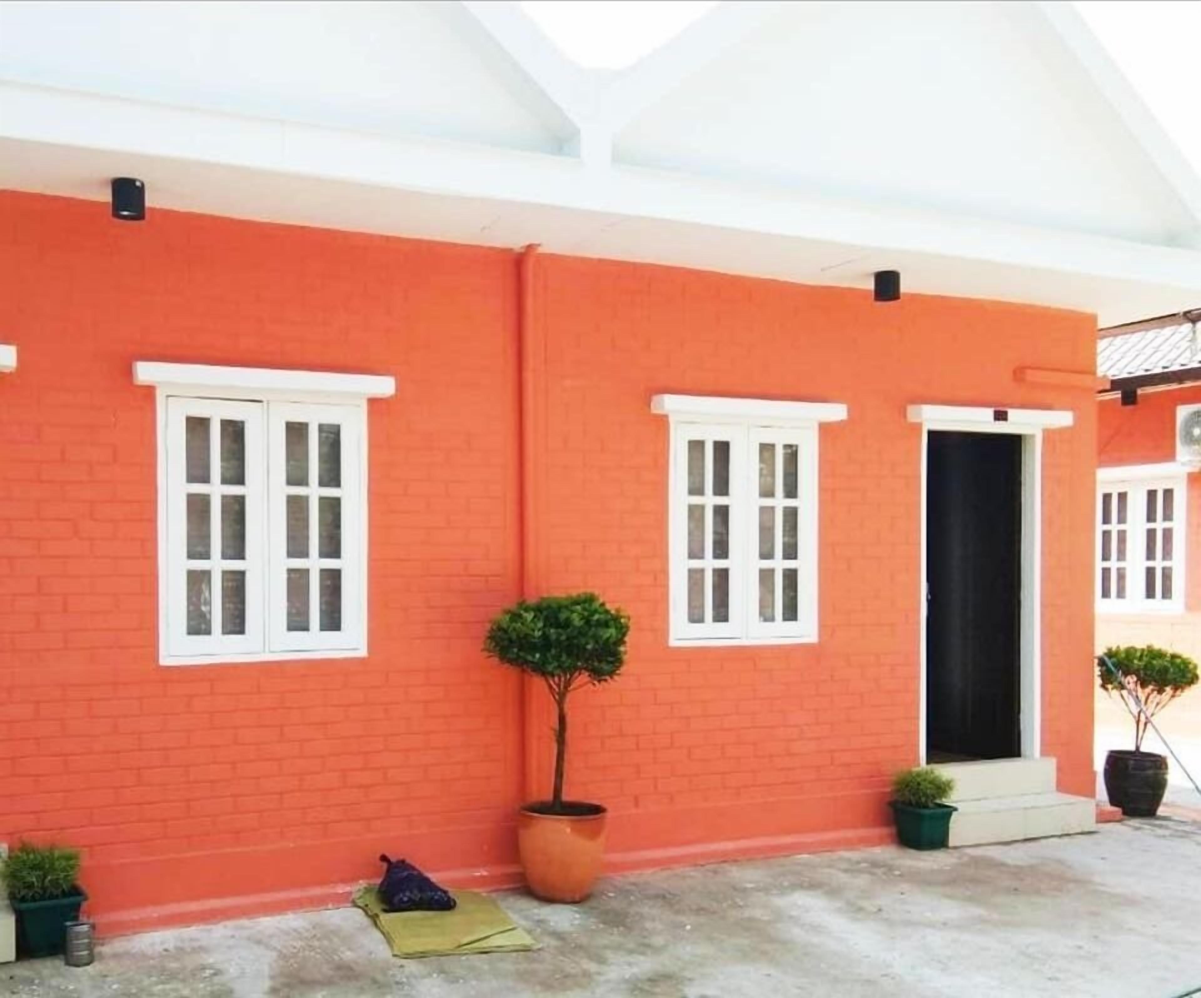 A two-story building with red brick walls and white-framed windows. There are two potted plants on the ground floor, one near the entrance and another by the windows.
