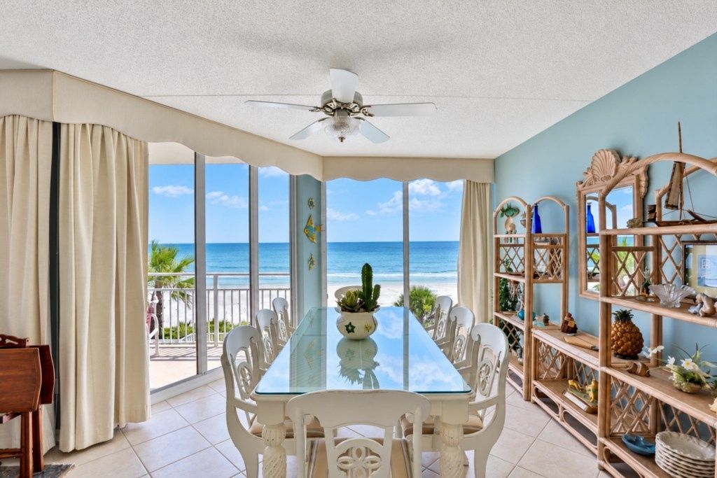 A dining area with a glass table and white chairs, overlooking a beach with a clear view of the ocean.