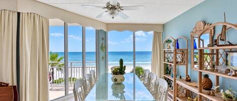 A dining area with a glass table and white chairs, overlooking a beach with a clear view of the ocean.