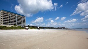 Ein Strand mit einem großen Hotel im Hintergrund, wolkenlosem Himmel und einigen Spaziergängern.