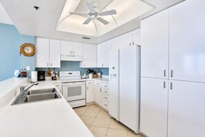 A modern kitchen with white cabinetry, a built-in oven, a refrigerator, and a ceiling fan. - Surf And Racquet 101 3 Bedroom Condo by RedAwning (New Smyrna Beach)