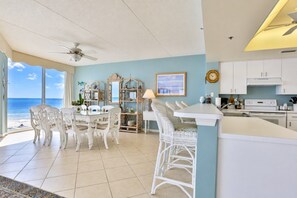 A dining area with a view of the ocean, a ceiling fan, and a kitchen with white cabinets. - Surf And Racquet 101 3 Bedroom Condo by RedAwning (New Smyrna Beach)