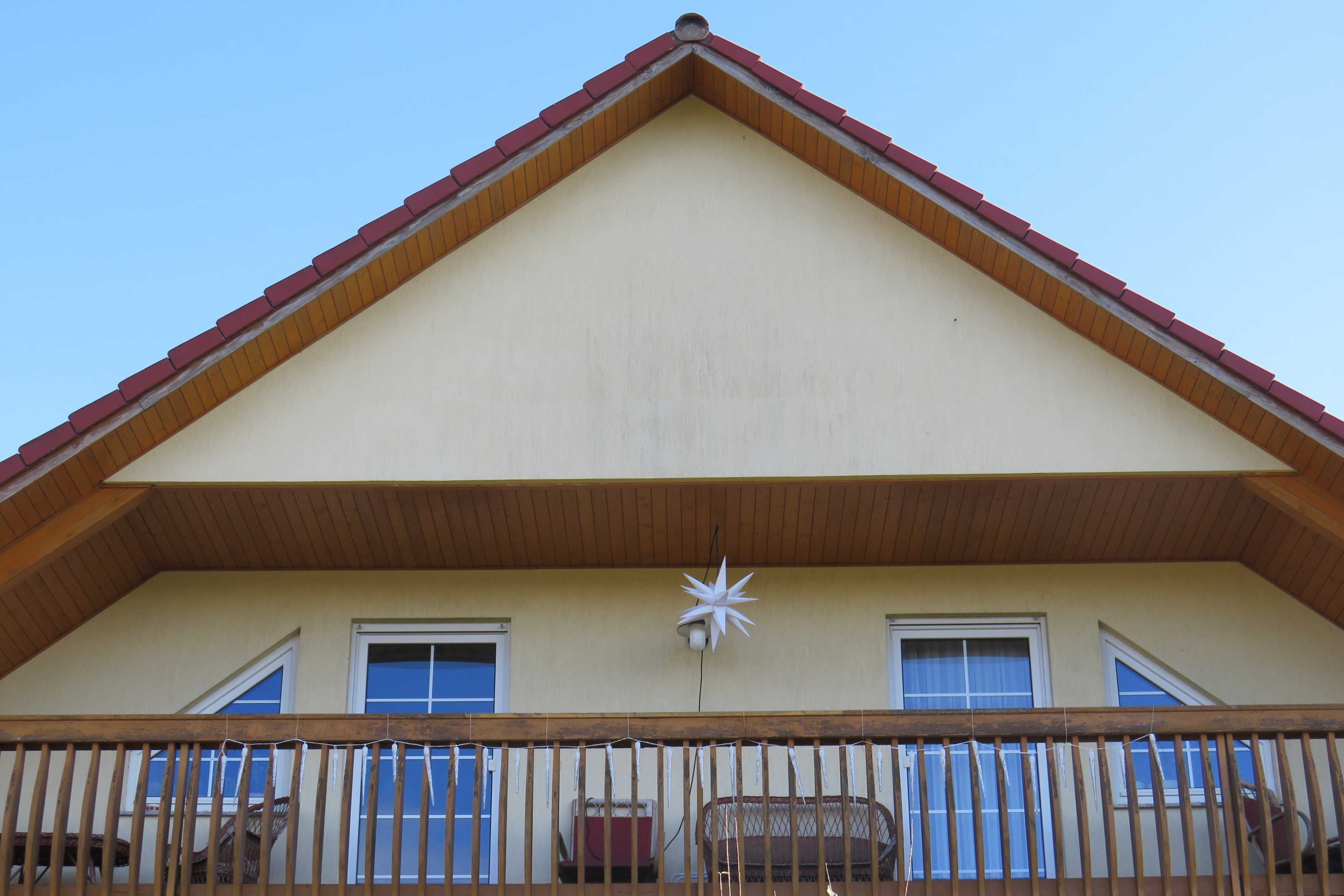 Un bâtiment doté d’un toit triangulaire, d’un balcon avec une balustrade en bois et de deux fenêtres ornées de rideaux bleus.