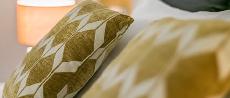 Et nærbillede af en seng med mønstrede puder og en lampe i baggrunden.