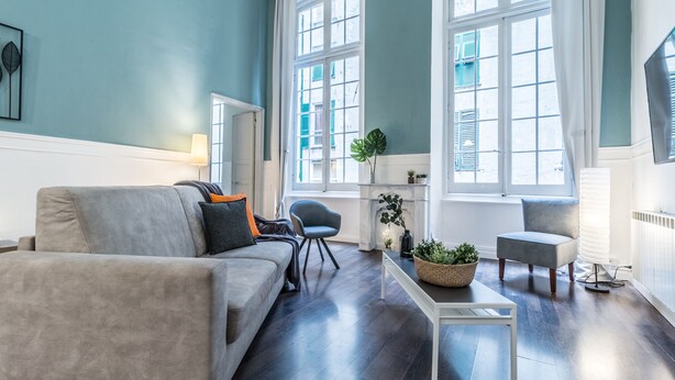 Una sala de estar moderna con un sofá gris, una mesita y una silla. Hay ventanas con marcos blancos y vistas a edificios.