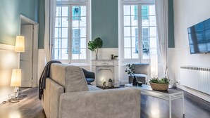 Una sala de estar moderna con un sofá, una mesa de centro y una chimenea. Hay dos ventanas con cortinas, un televisor de pantalla plana montado en la pared y una puerta que da a otra habitación.
