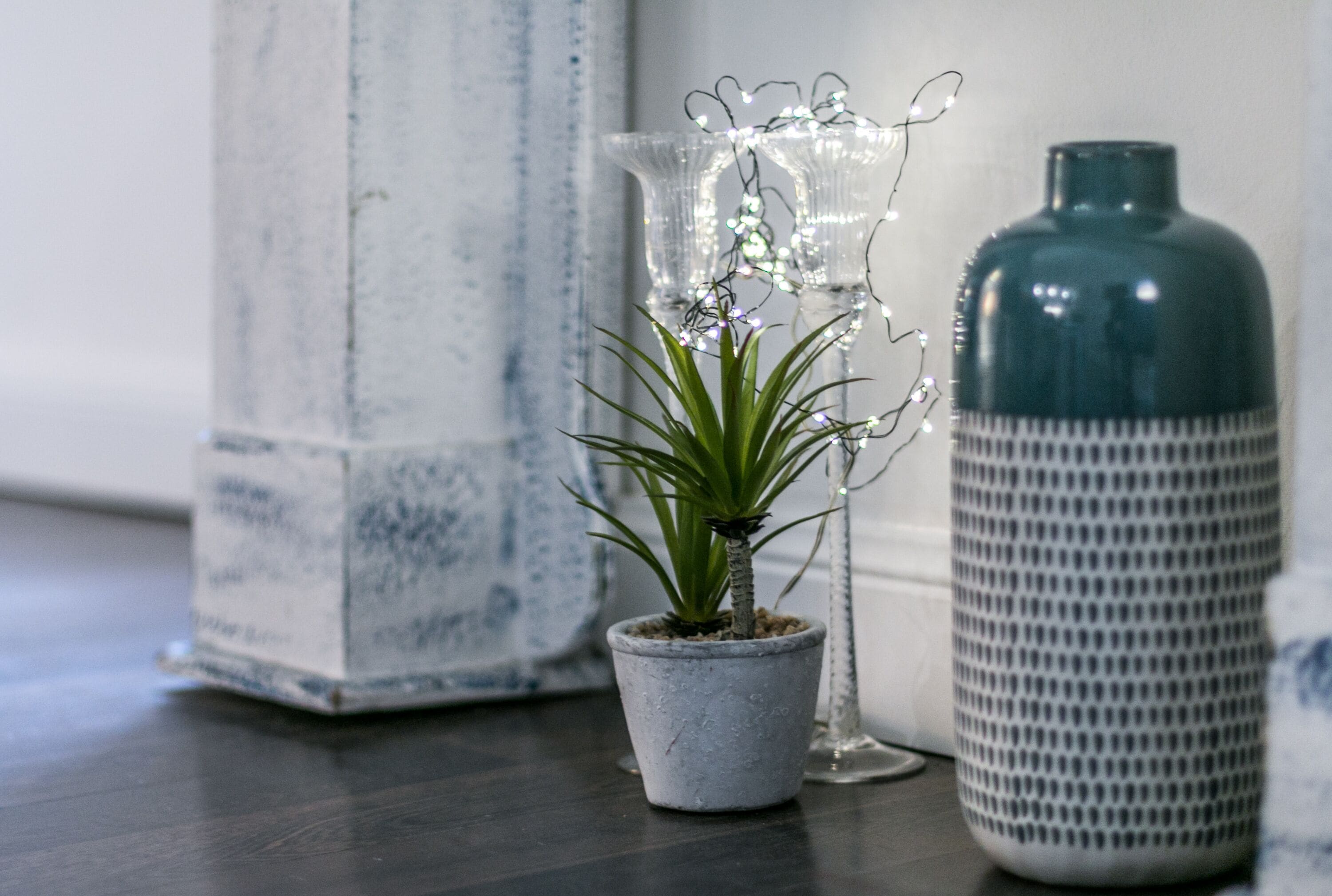 A small potted plant, a clear glass vase with lights, and a textured ceramic container on a wooden surface.