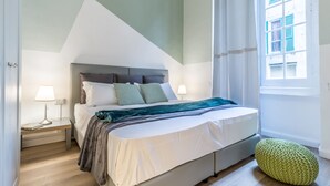Un dormitorio con cama, mesitas de noche, lámpara, ventana y un otoman verde.