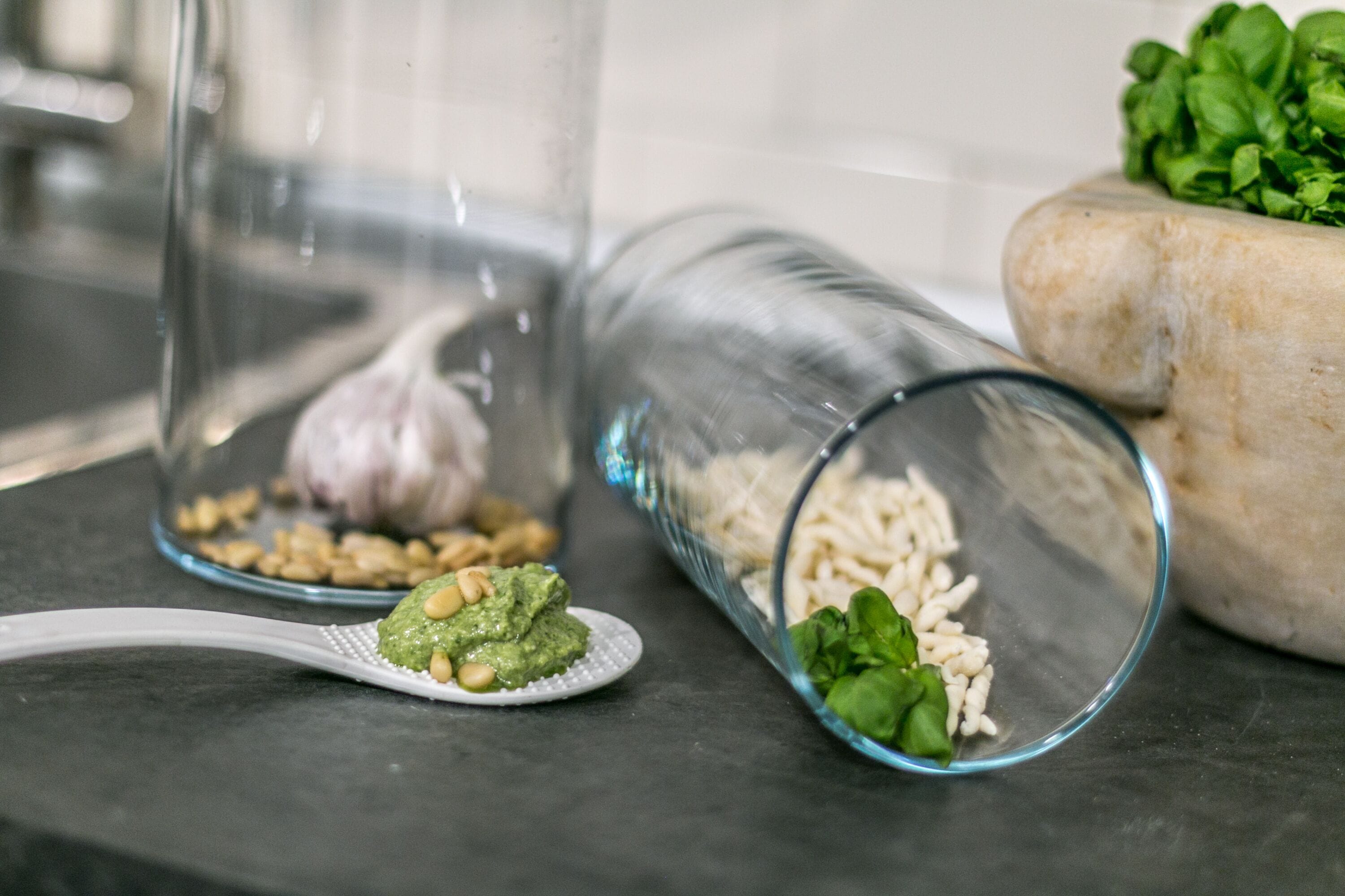 Een aanrecht met een glazen pot met rijst, een lepel met groene saus, en een vijzel met groene bladeren.