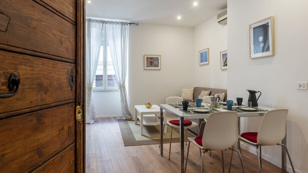 A dining area with a wooden cabinet, a white table, and chairs. There are framed pictures on the wall and a window with curtains. - Via degli Avignonesi (Rome)