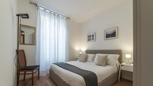 A bedroom with a bed, bedside tables, a chair, a desk, a lamp, a mirror, and a window with curtains. - Via degli Avignonesi (Rome)