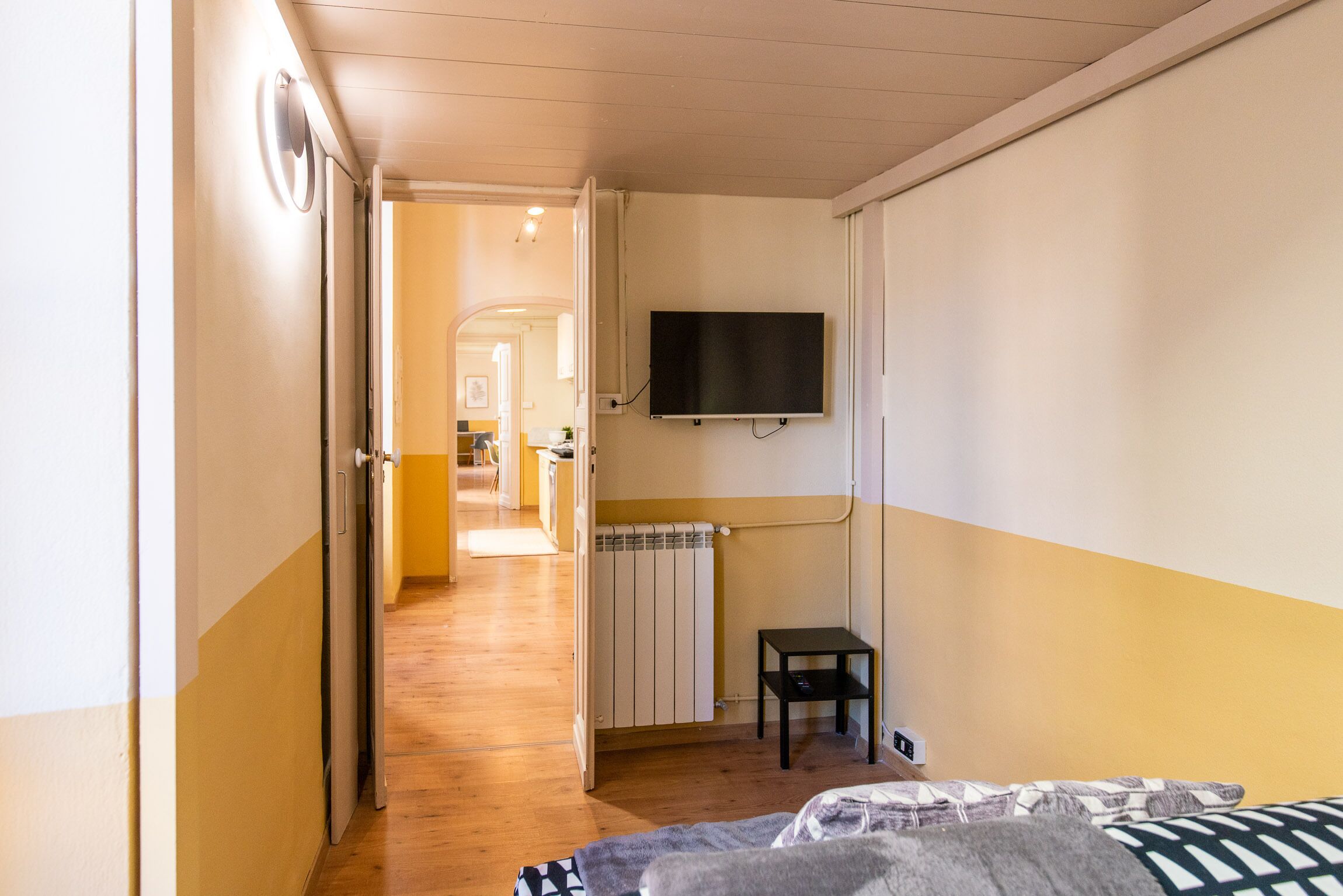 Ein Zimmer mit einem Bett, einem an der Wand befestigten Fernseher, einem Heizkörper und einem kleinen Tisch.