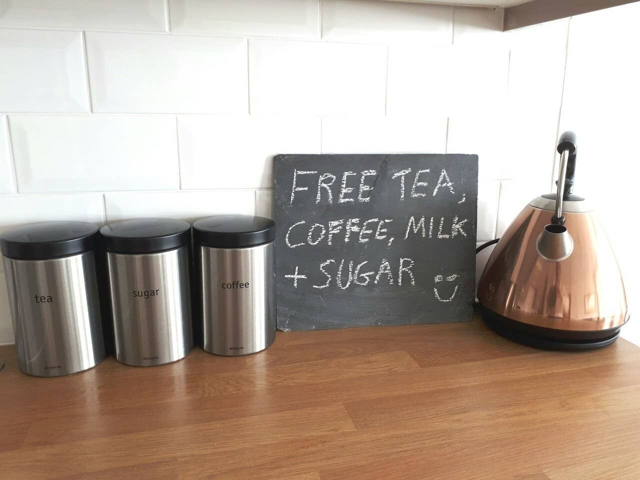 Stainless steel containers labeled tea, sugar, and coffee, a copper kettle, and a chalkboard sign offering free tea, coffee, milk, and sugar.
