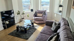 A living room with a flat-screen TV, a glass coffee table, a purple sofa, and two windows with blinds. - River views By Royal William Yard (Plymouth)