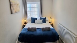A narrow hotel room with a large bed, two bedside lamps, a radiator, and a window with curtains. - River views By Royal William Yard (Plymouth)