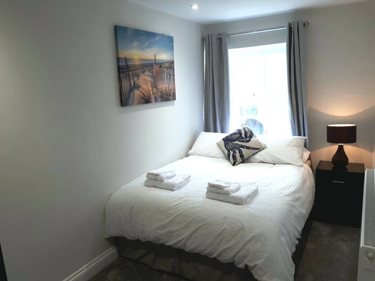 Ein ordentlich bezogenes Bett mit weißen Leinen und Kissen, ein Nachttisch mit Lampe sowie ein Bild an der Wand.