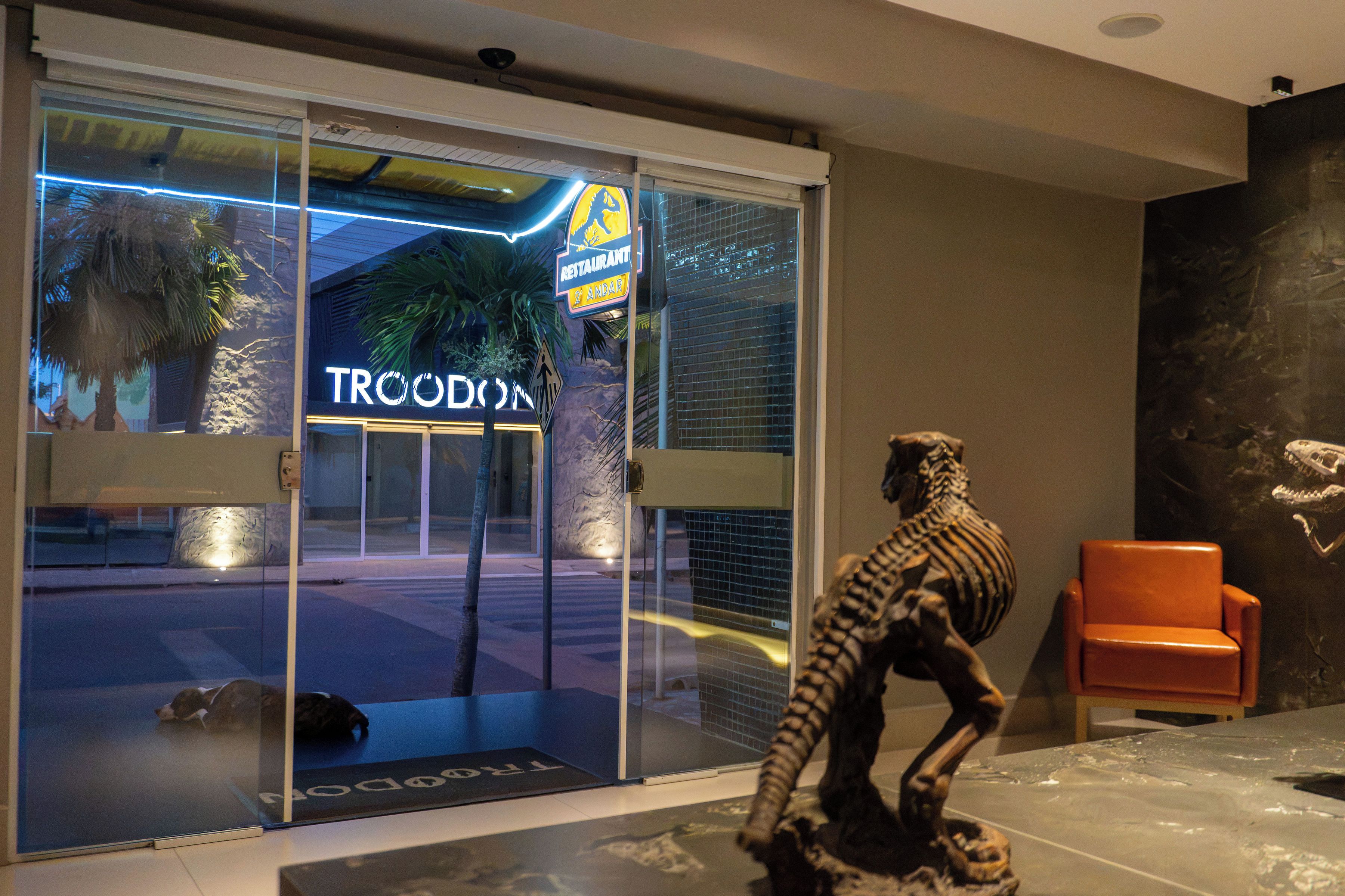 Entrada de hotel con puerta de vidrio, un letrero que dice 'TRCODON' y una estatua de dinosaurio.