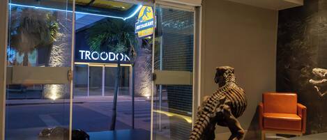 Entrada de hotel con puerta de vidrio, un letrero que dice 'TRCODON' y una estatua de dinosaurio.