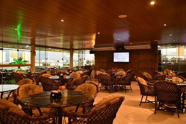 An indoor dining area with wicker chairs and tables, a large window with a view of buildings, and an air conditioning unit on the wall. - Troodon Park Hotel (Sousa)