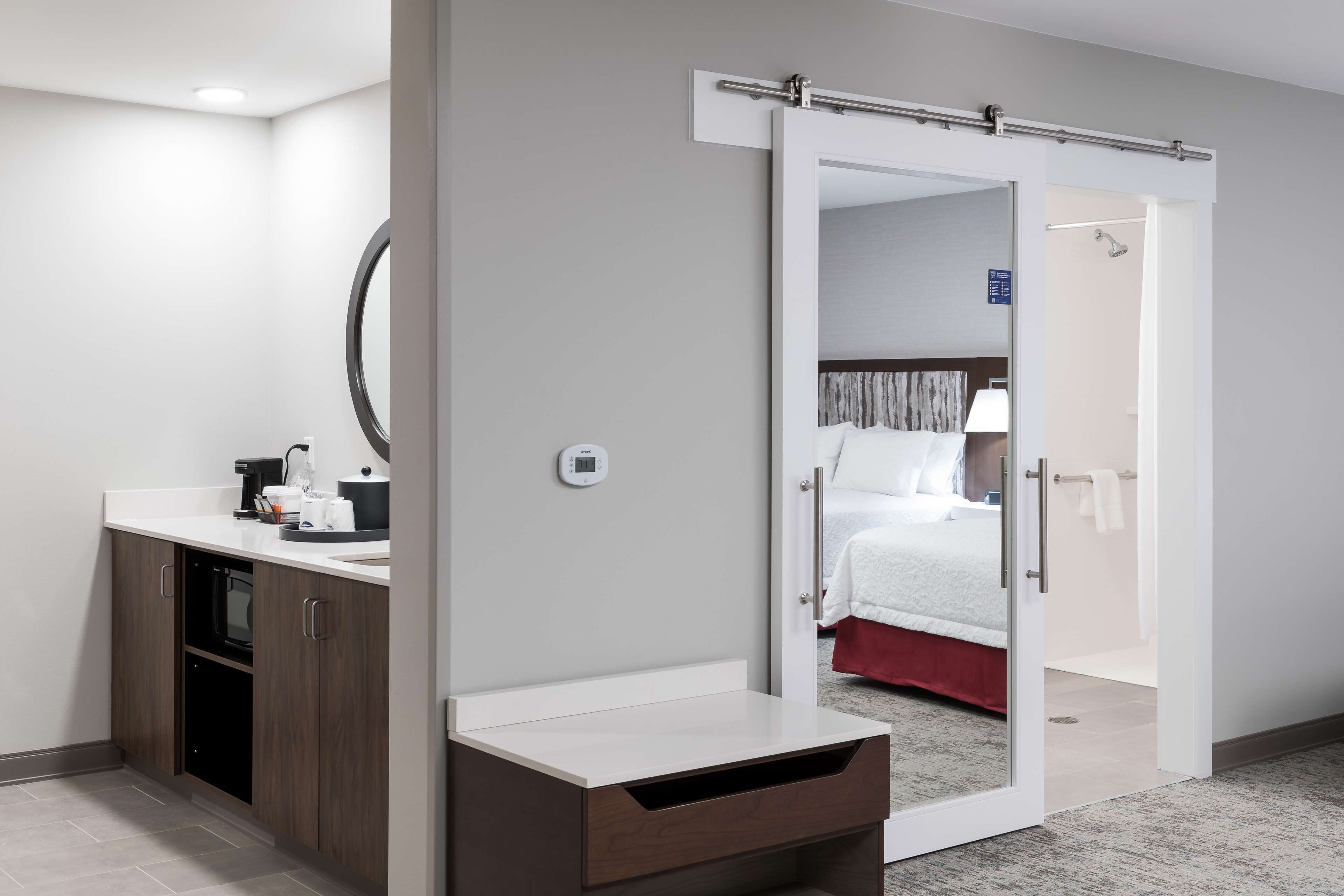 A hotel room with a kitchenette, a mirror, a sliding door to a bedroom, and a small trash bin.