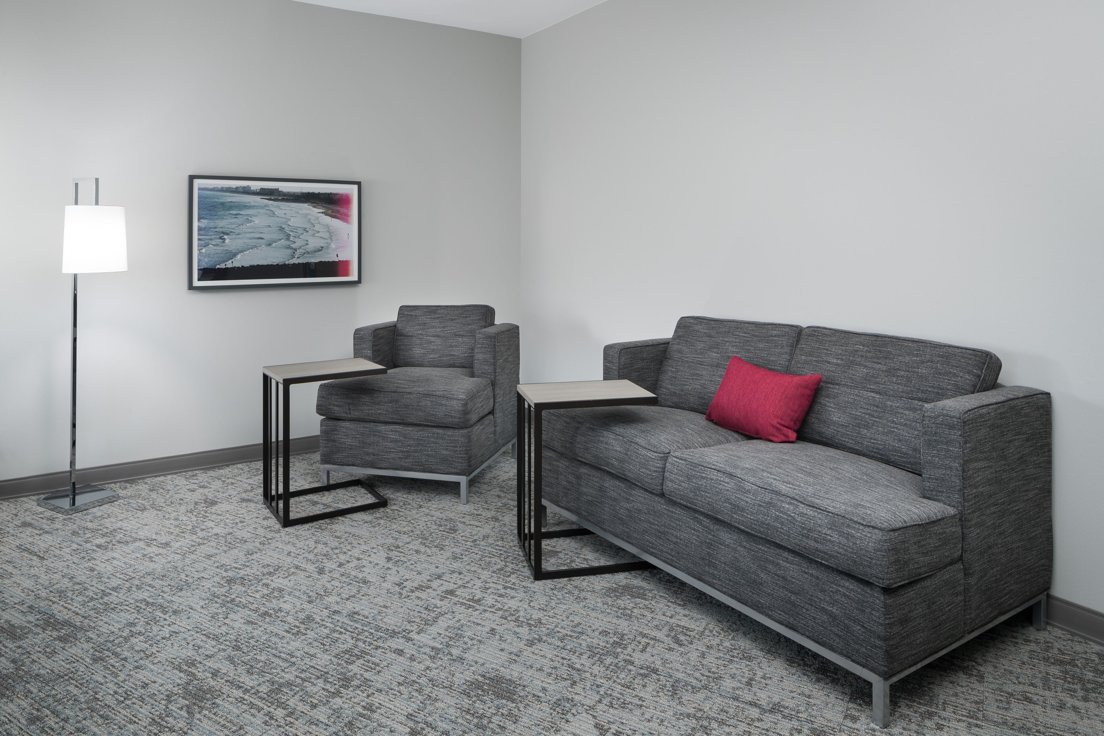 Una sala de estar moderna con un sofá gris, un sillón más pequeño y una mesita auxiliar. Un cuadro enmarcado cuelga de la pared.