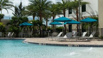 A pool area with lounge chairs and umbrellas, surrounded by palm trees and a building.