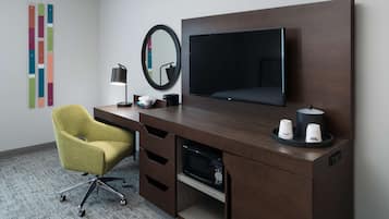 A hotel room with a wooden desk, a television, a mirror, a yellow chair, and a coffee machine.