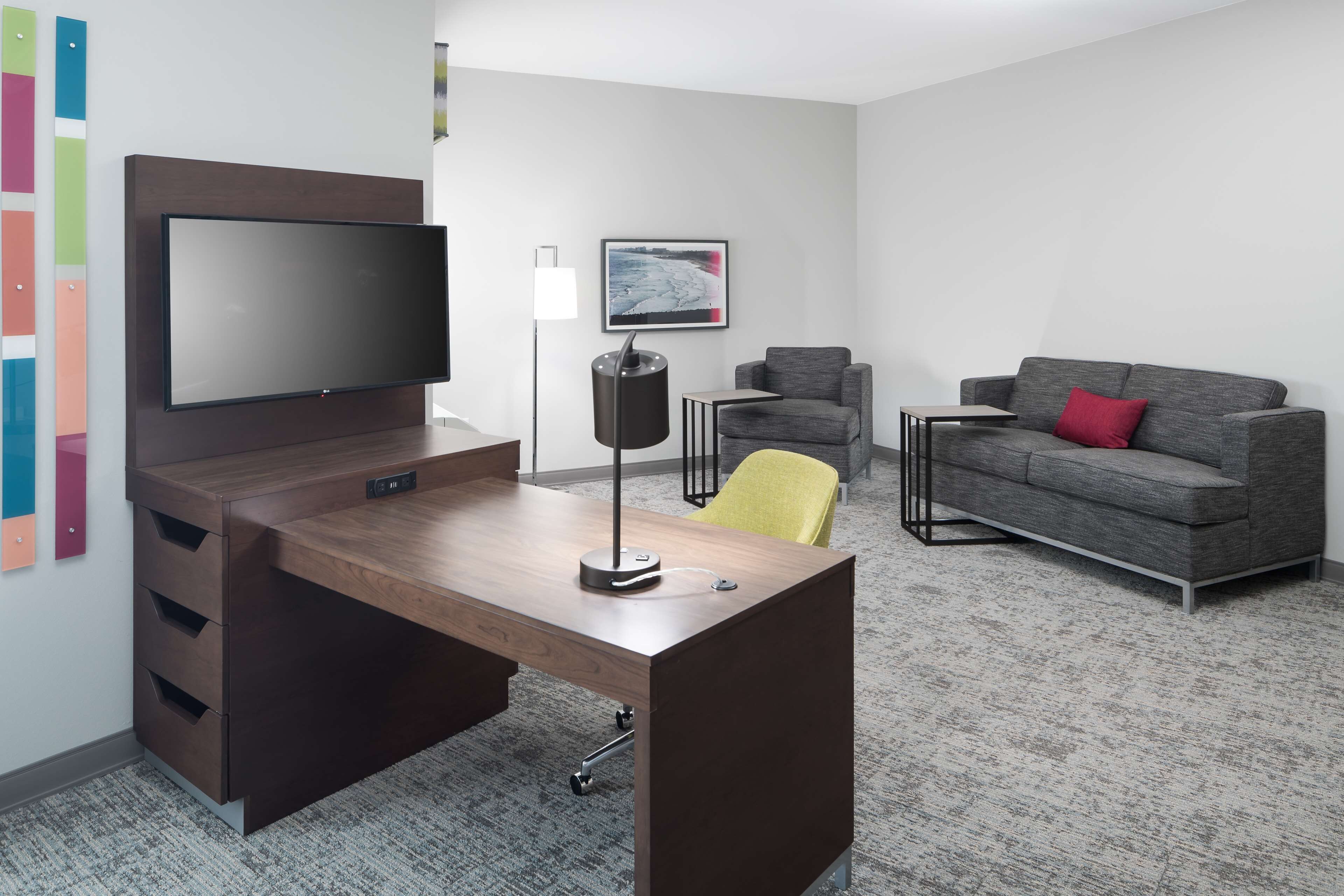 Habitación de hotel moderna con un escritorio de madera, un televisor de pantalla plana, un sofá gris con un cojín rojo, una silla amarilla y un cuadro en la pared.