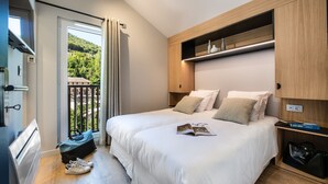 A modern hotel room with a large bed, a wooden headboard, a built-in shelf, and a view of a balcony with greenery.
