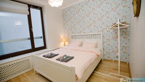 A bedroom with a bed, a window, a radiator, and a wall with floral wallpaper. - Warsaw Concierge Vistula Apartment (Warsaw)