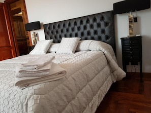 Ein ordentlich bezogenes Bett mit einem gesteppten Kopfteil, ein Stapel gefalteter Handtücher und ein Nachttisch mit einer Lampe.
