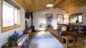 A cozy living room with a wooden ceiling, a brown leather sofa, a wooden coffee table, and a dining area with a table and chairs. - Laurel Lodge - Log Cabin - St Florence (Tenby)