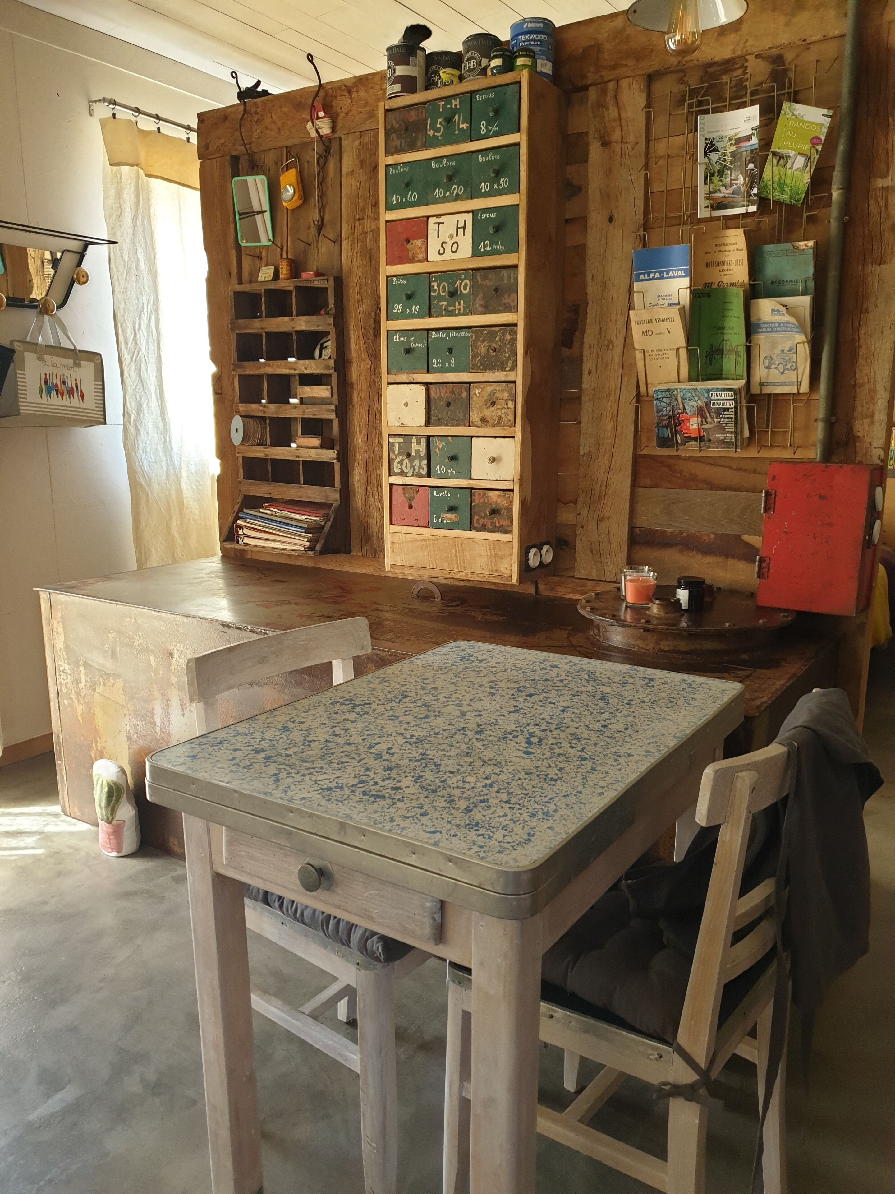 A rustic room with a wooden table and chairs, a shelf with vintage items, and a wall with various objects and papers.