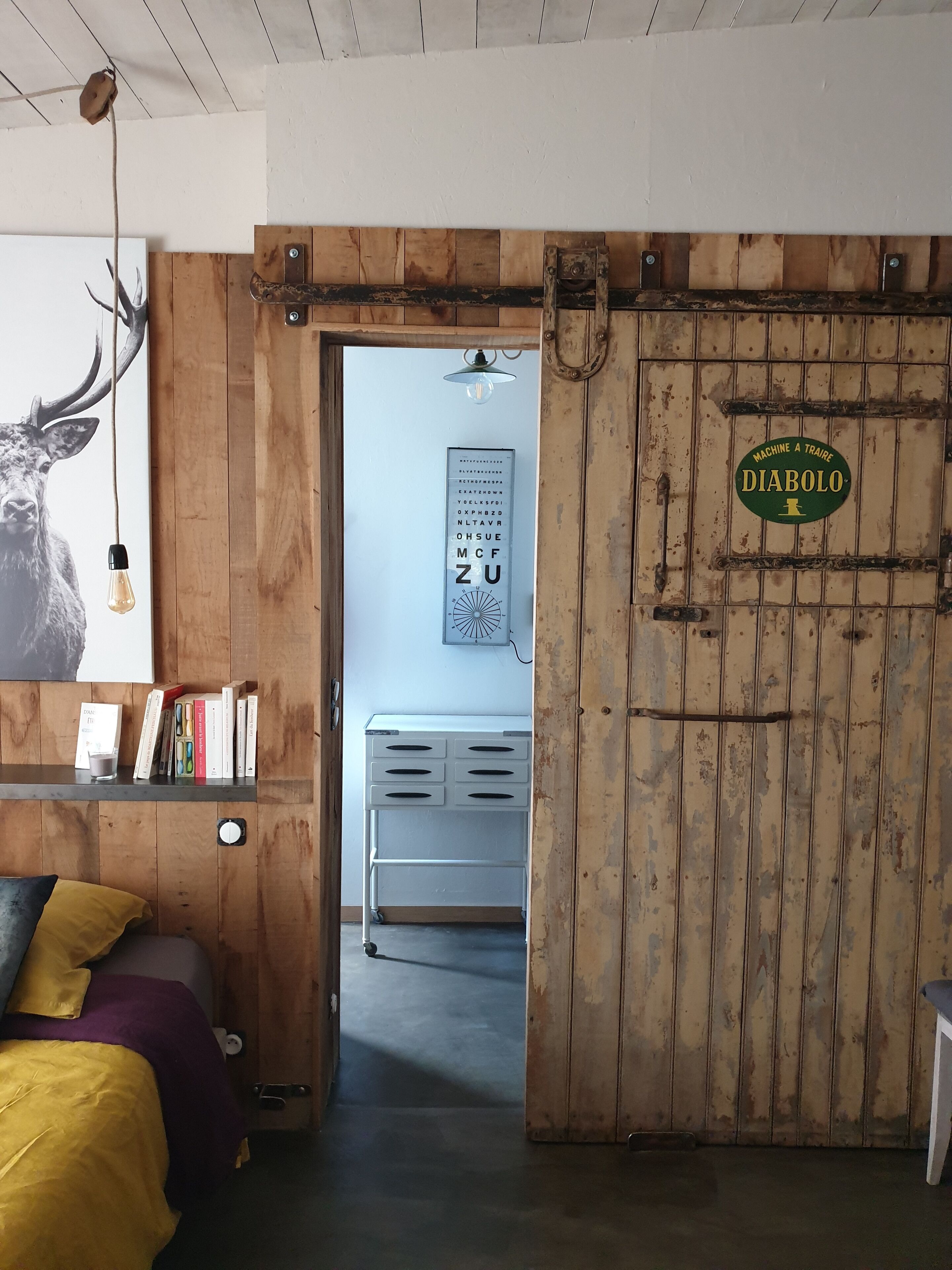 A room with a wooden door labeled "DIABOLO," a bookshelf with books, a framed poster, and a bed with a yellow and purple blanket.