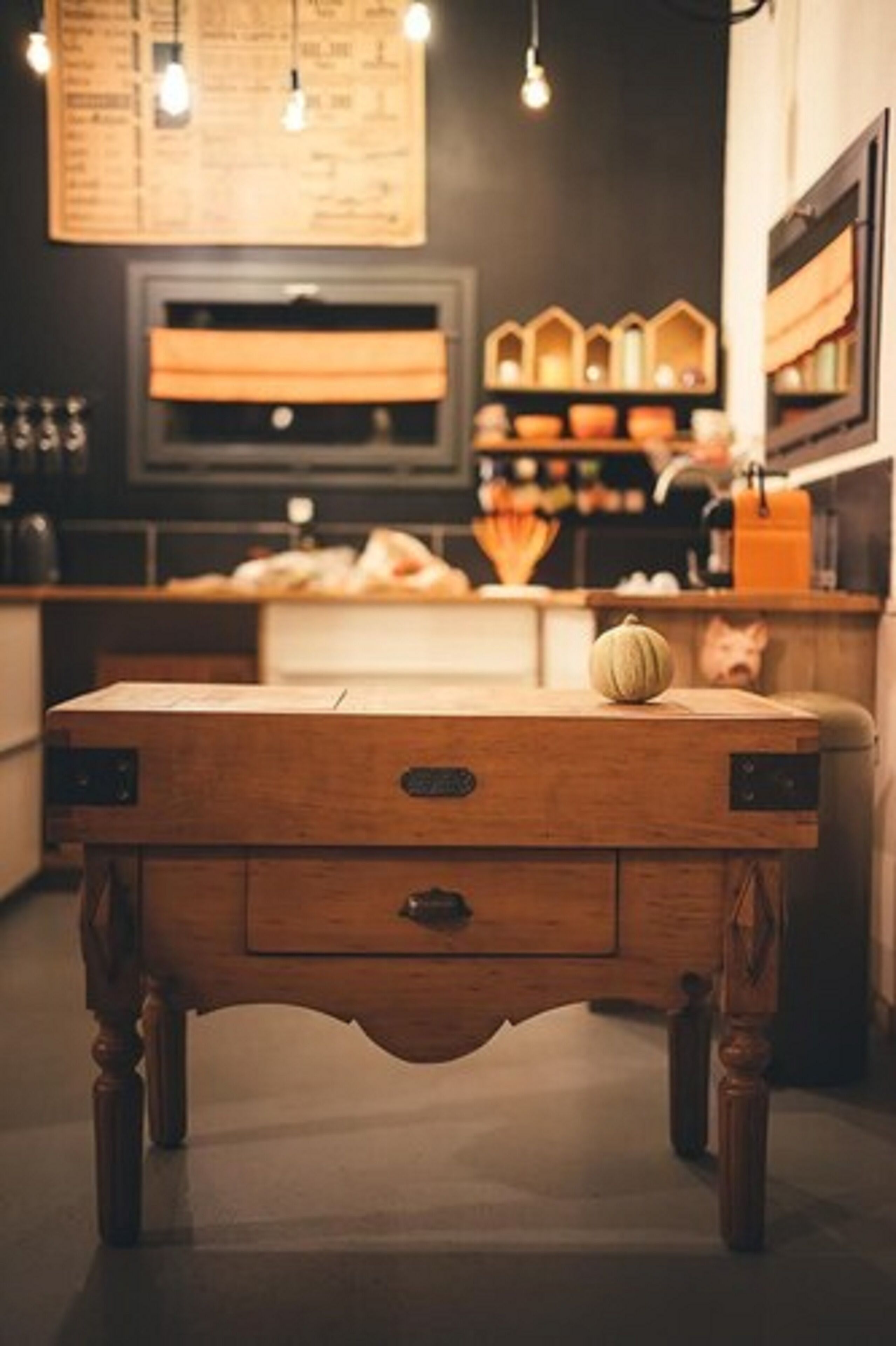 Una mesa de madera con un cajón, una calabaza y un cerdito alcancía en una cocina acogedora, con luces colgantes y estantes de madera al fondo.