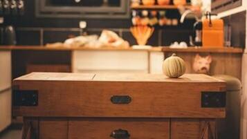 Una mesa de madera con un cajón, una calabaza y un cerdito alcancía en una cocina acogedora, con luces colgantes y estantes de madera al fondo.