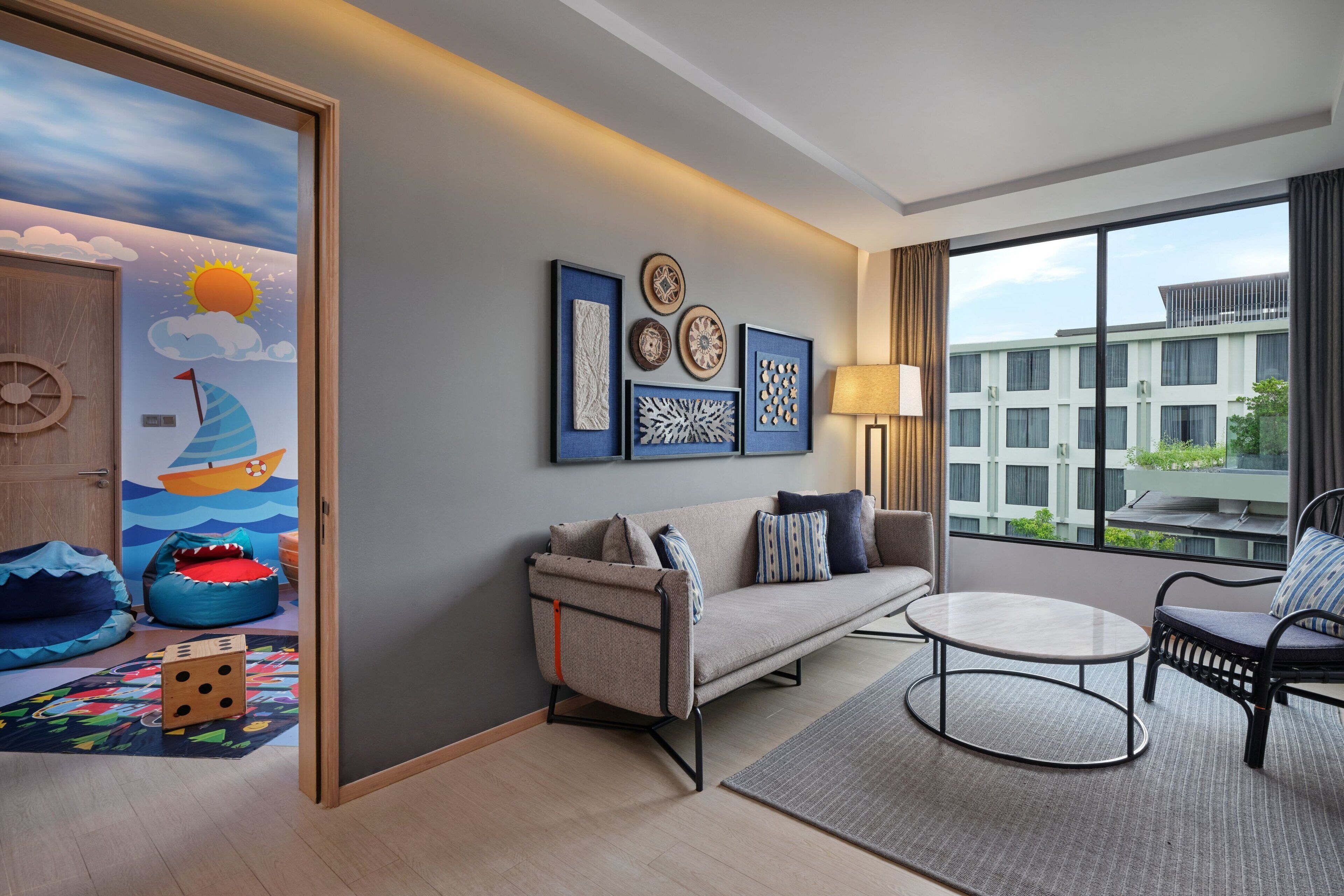 A modern hotel room with a sofa, armchair, and a round table. There is a large window offering a view of the building.