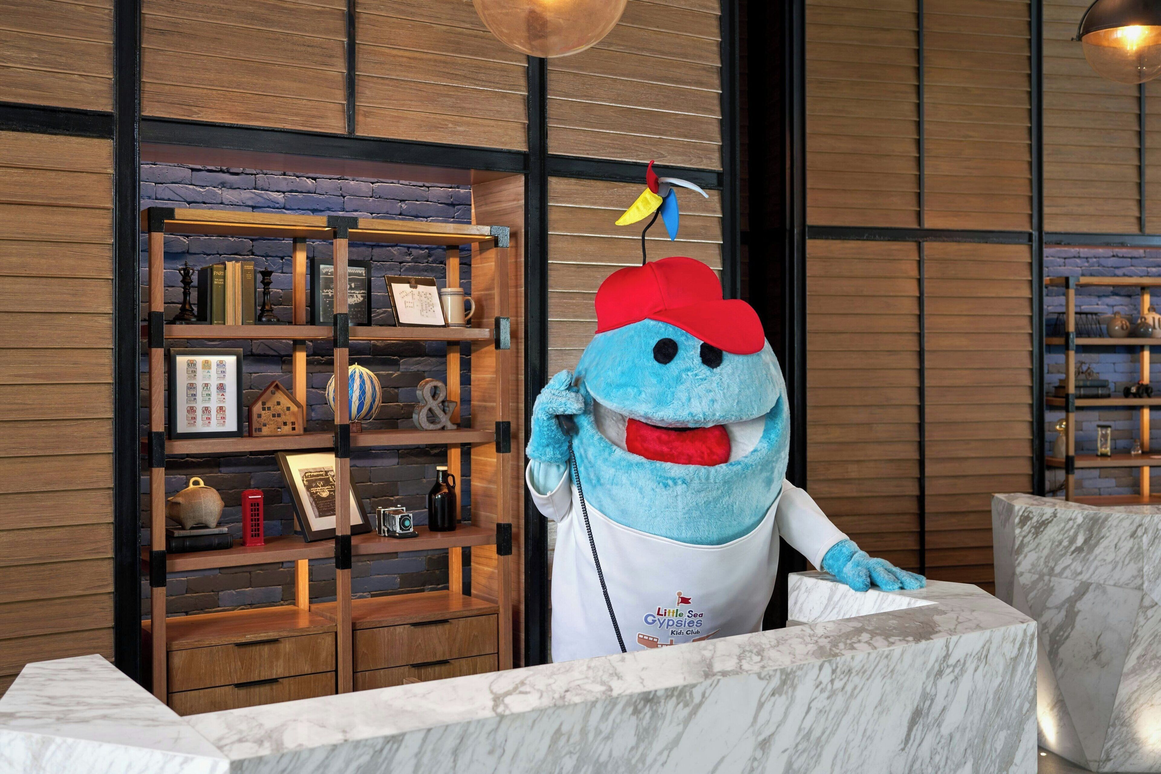 Een mascotte in een witte koksoutfit met een rode hoed, staand voor een houten boekenkast met diverse objecten.
