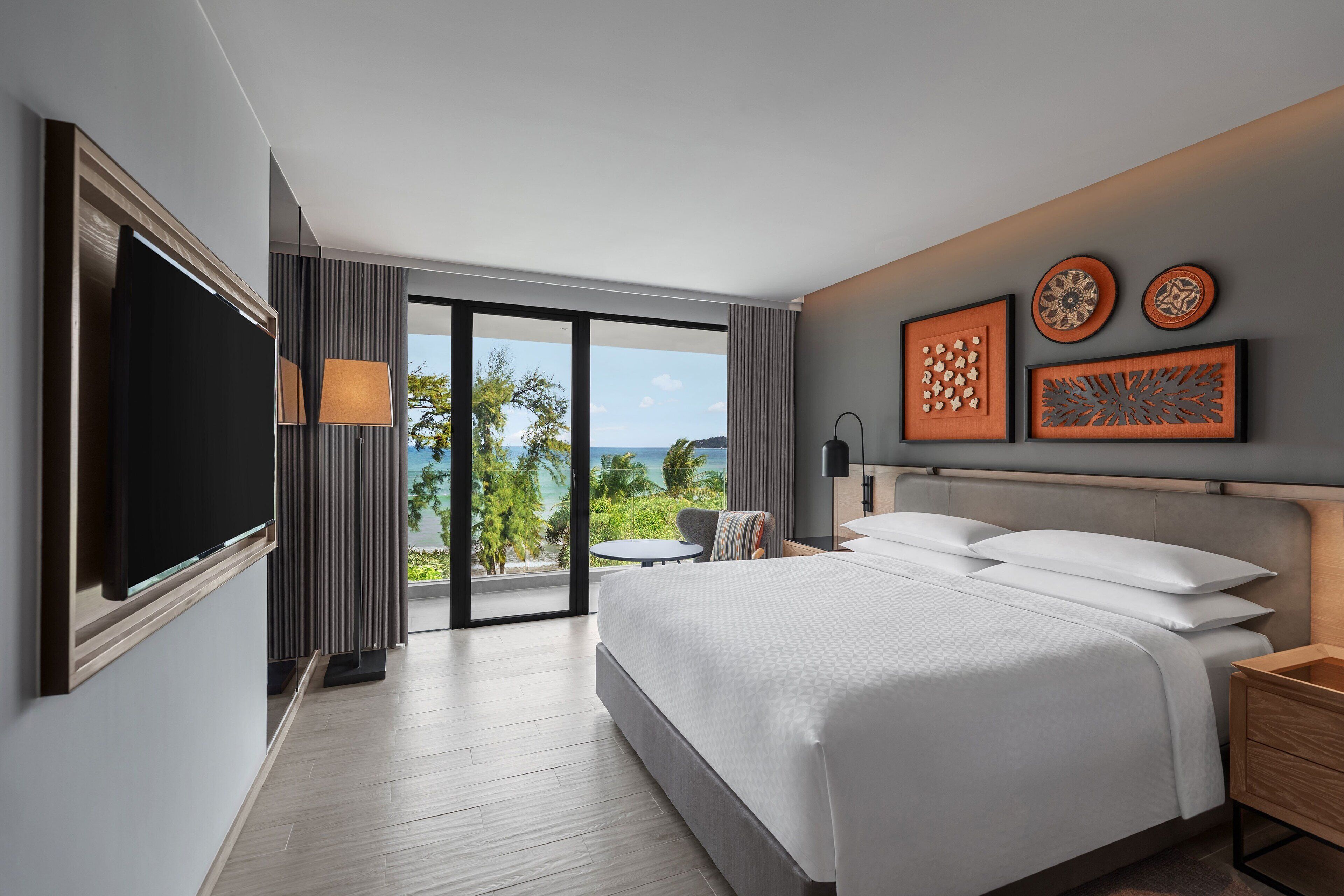 A modern hotel room with a large bed, a flat-screen TV, a balcony with a view of the ocean, and artwork on the walls.