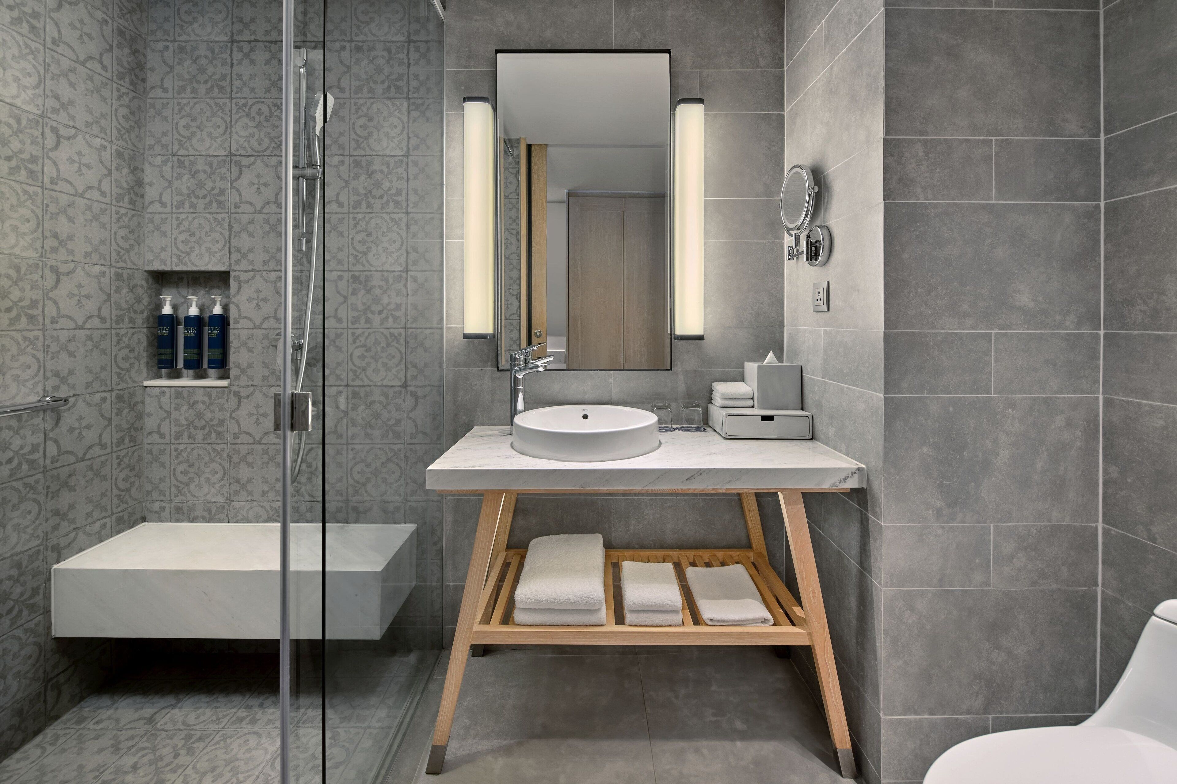 Une salle de bain moderne dotée d’une douche à parois de verre, d’un meuble-lavabo en bois avec un vasque blanche et d’une étagère intégrée pour les articles de toilette.