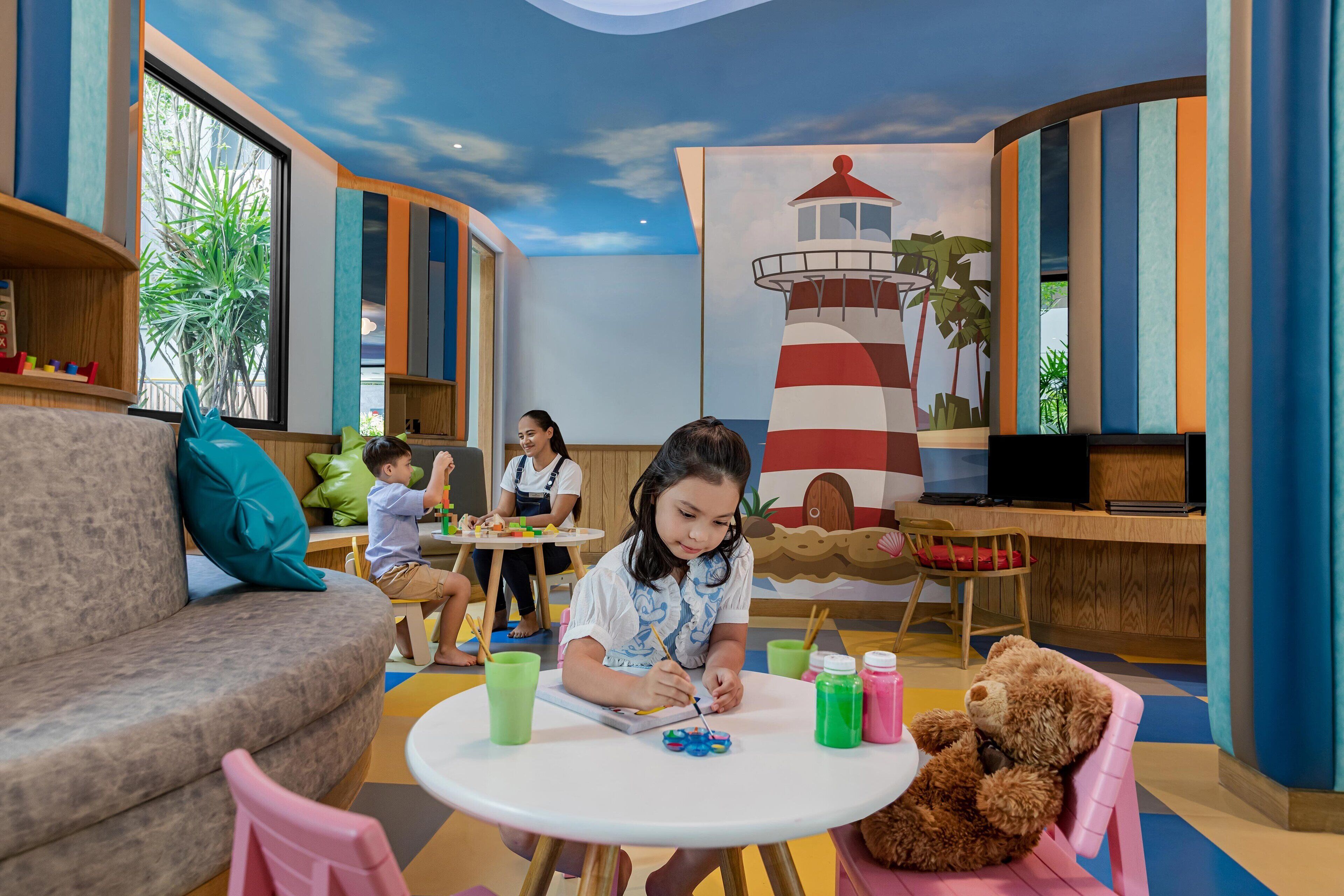 A children's play area with a large mural of a lighthouse, a sofa, a table with art supplies, and a teddy bear.