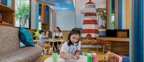 A children's play area with a large mural of a lighthouse, a sofa, a table with art supplies, and a teddy bear.
