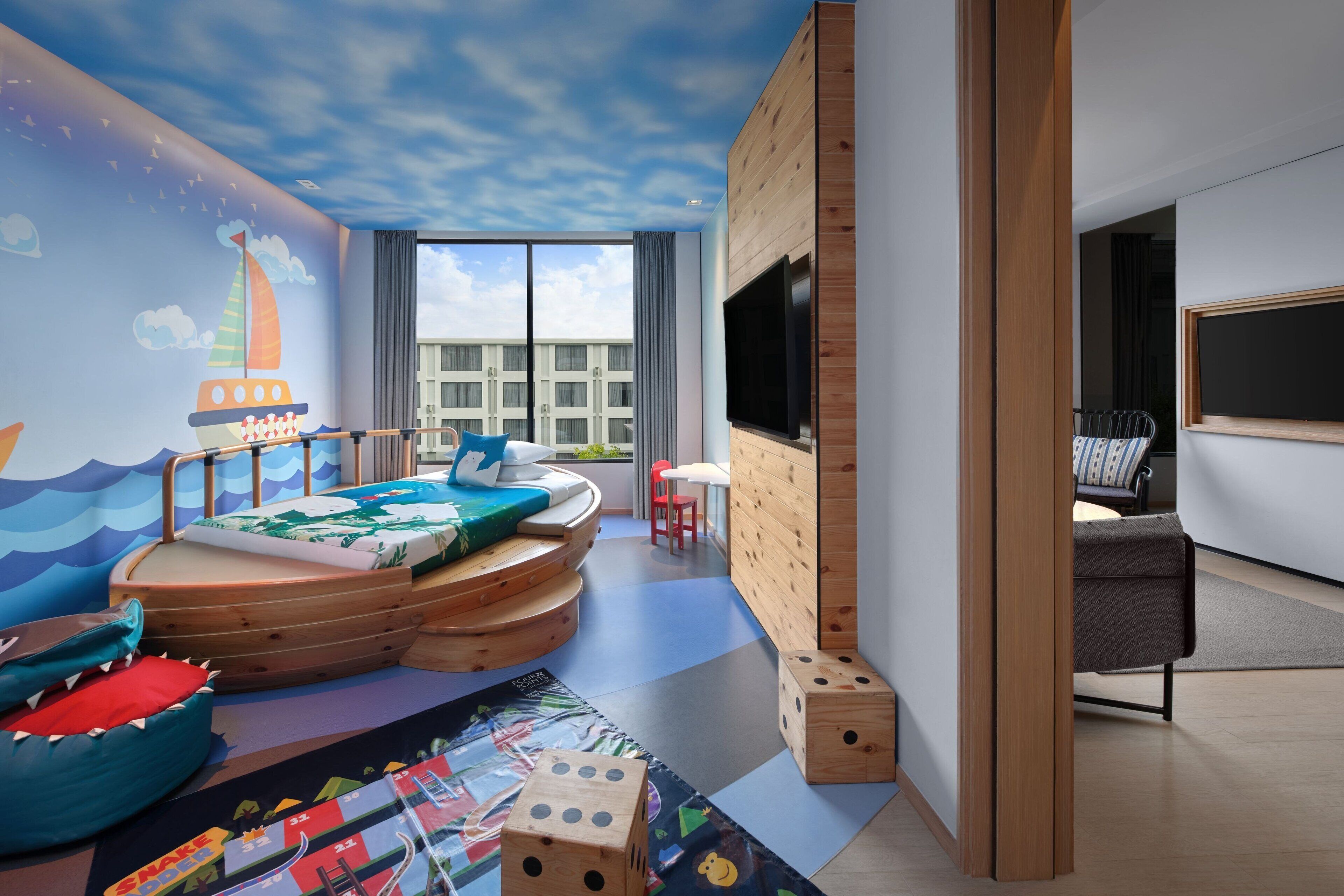 Ein Spielzimmer für Kinder mit Wandgemälde eines Segelboots, einem Spielbereich inklusive Rutsche und einem großen Fenster mit Blick auf die Stadt.