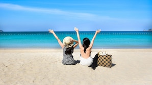 Two people sitting on a sandy beach with their arms raised, facing a calm sea and clear sky.