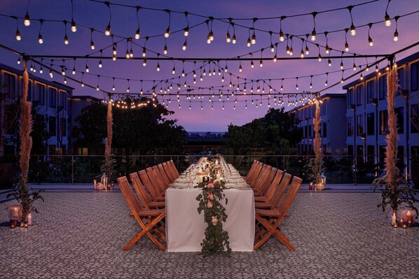 Un área de comedor al aire libre con una larga mesa dispuesta para una comida, rodeada de sillas, decorada con luces colgantes y velas.