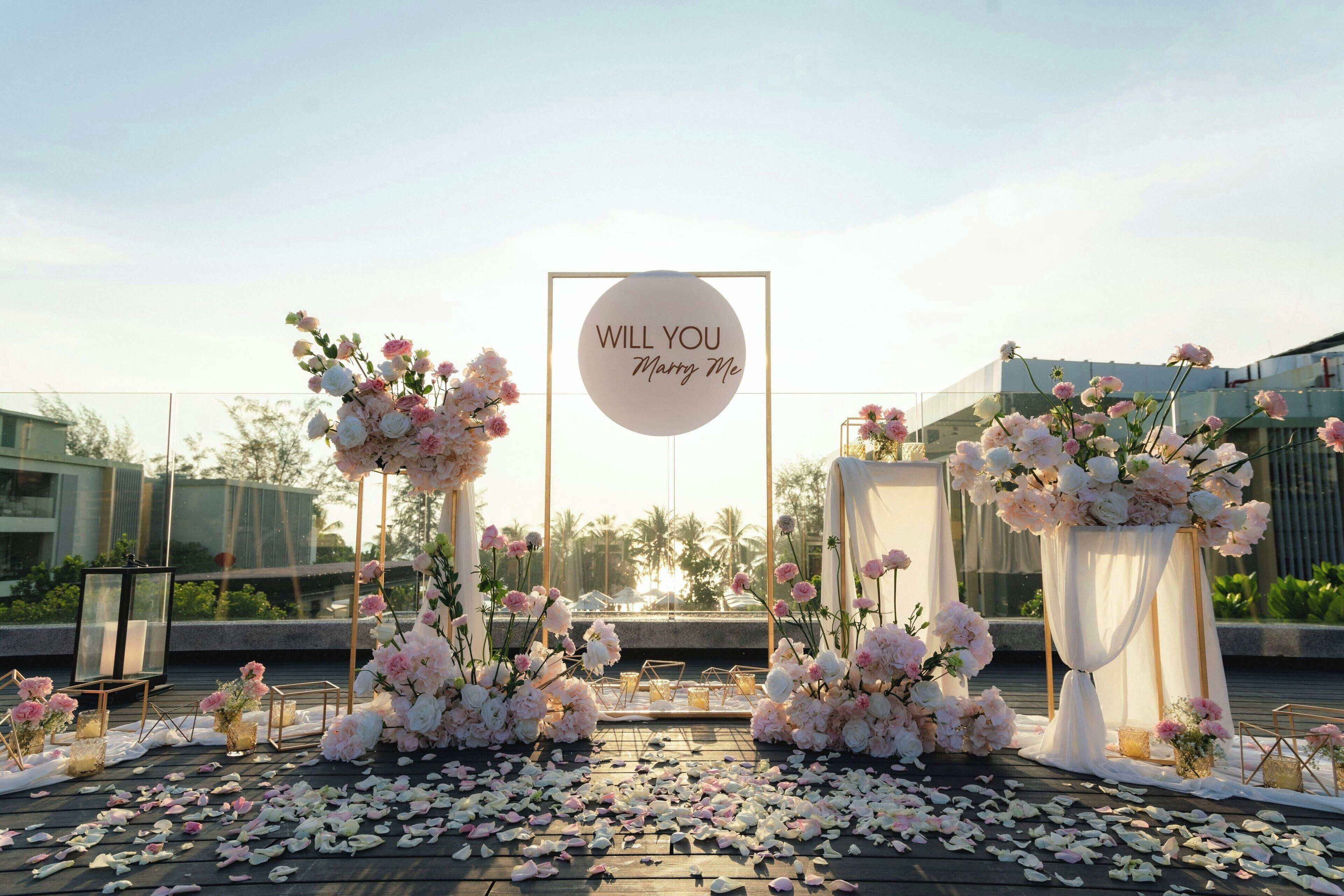 A wedding ceremony setup with a 'WILL YOU marry me' sign, floral arrangements, and a wooden deck.