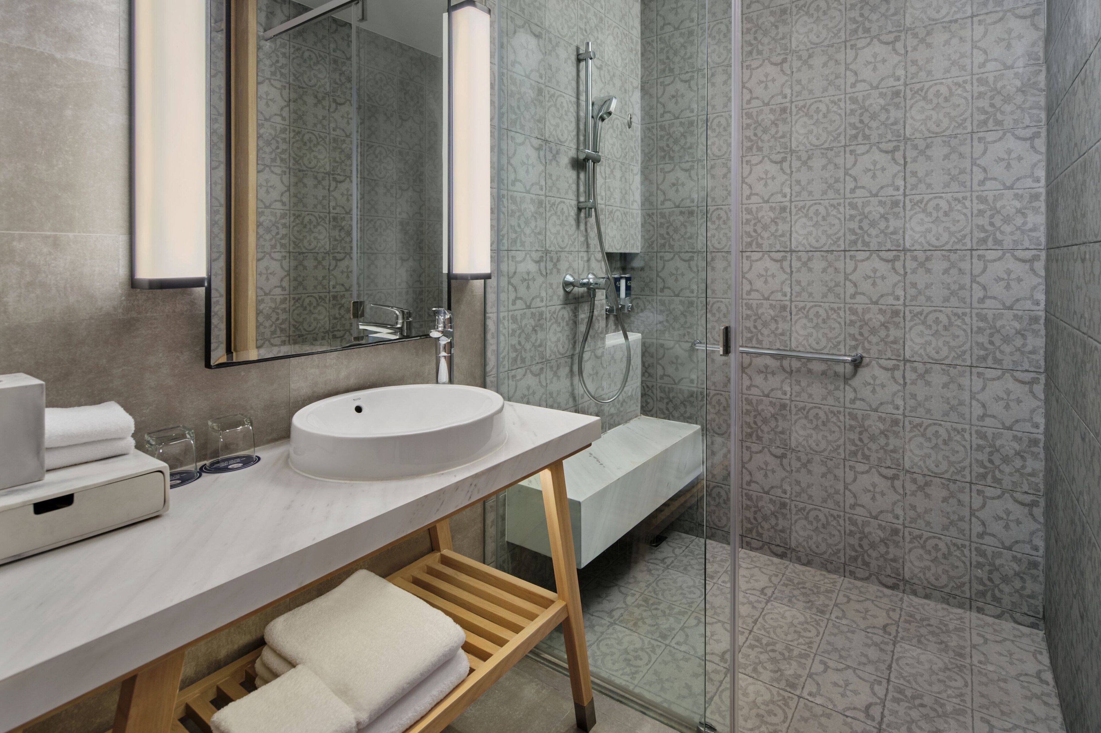 A modern bathroom with a large mirror, a round sink, a glass shower enclosure, and a wooden shelf with towels.