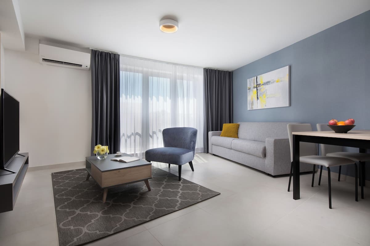 Ein modernes Wohnzimmer mit Flachbildfernseher, grauem Sofa, Esstisch mit Stühlen und einem großen Fenster mit Vorhängen.